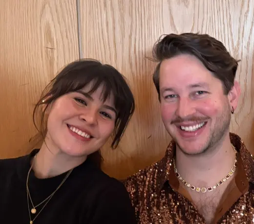 Two coworkers smiling together in front of a wood-paneled wall.