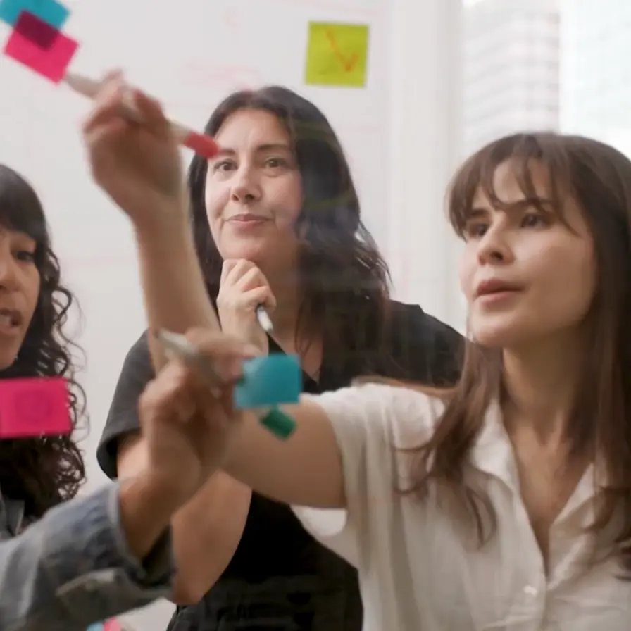 Three female coworkers brainstorm together, writing and placing sticky notes on a glass board in a bright, modern office environment.