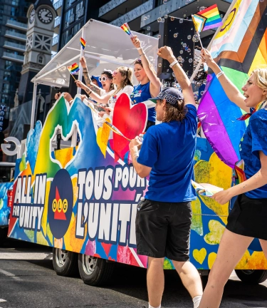 A colourful OLG parade float with rainbow flags and cheering people celebrates Pride on a city street, surrounded by tall buildings.