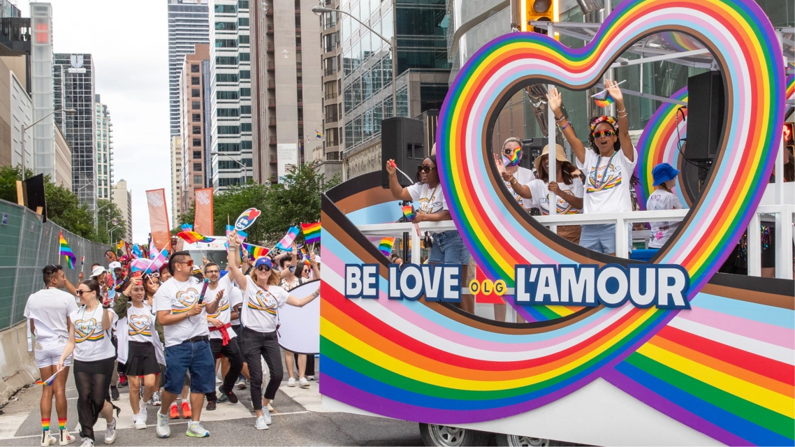 A colorful Pride parade float with a heart frame and the words Be Love and L’Amour, surrounded by people celebrating and waving flags.