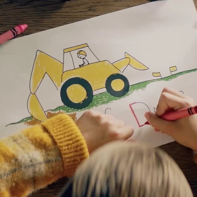 A child colours a drawing of a yellow excavator on green grass with a stick figure operator. The child is wearing a yellow sweater.
