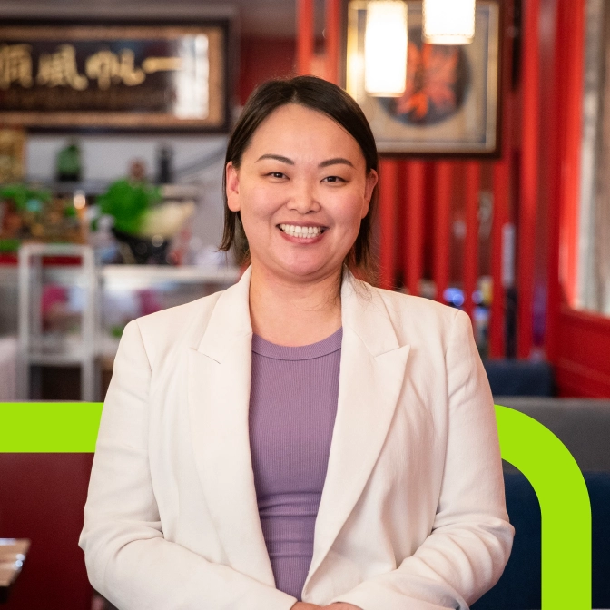 A woman with brown hair, wearing a white blazer and purple top, smiles while standing in a restaurant with red walls and Asian-inspired decor.