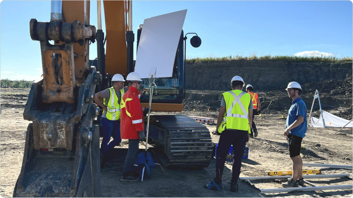 Outdoor filmset with a long arm excavator and people in hard hats and safety vests